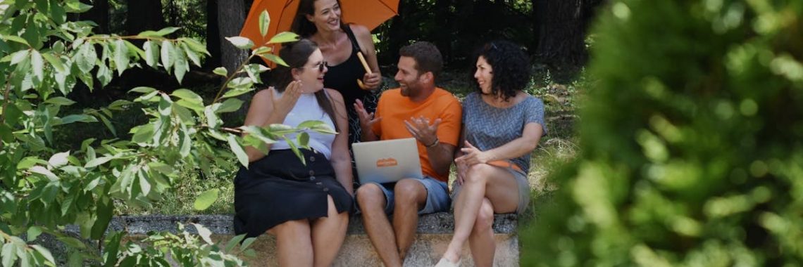 A diverse group of adults enjoying a conversation in nature, sitting with a laptop and umbrella on a sunny day.