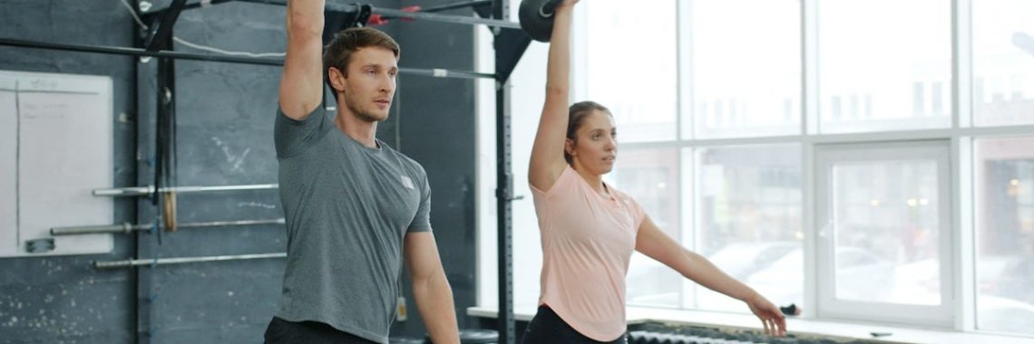 Young couple man and woman are training in gym weightlifting squatting with kettlebells together concentrated on bodybuilding exercises indoors.